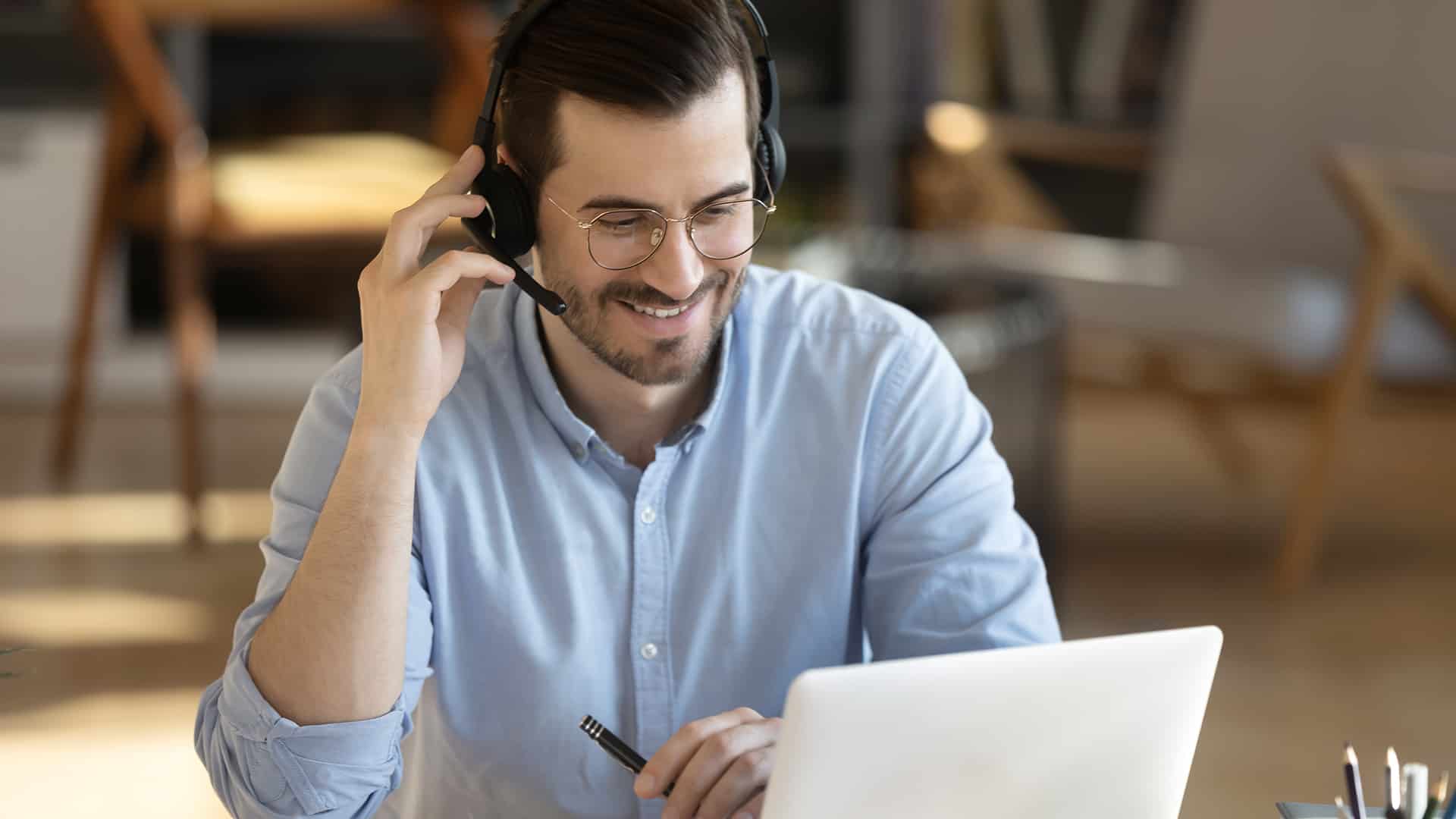 Smiling millennial Caucasian man in headset look at laptop screen work online from home office. Happy young man in headphones watch webinar take course or training on web. Distant education concept.