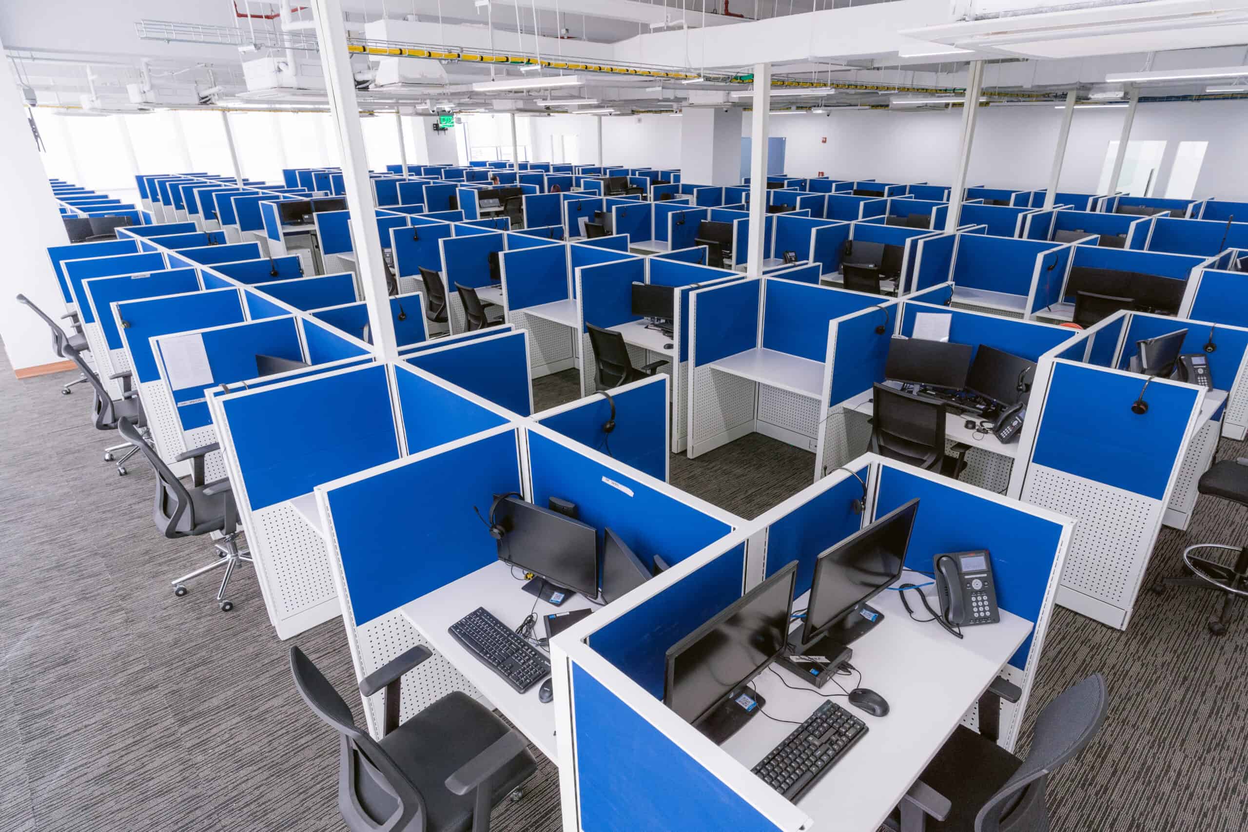 An empty NEARSOL Production Area featuring numerous workstations separated by blue dividers. Each cubicle is equipped with dual monitors, keyboards, telephones, and headsets, arranged in a neat and organized manner. The workspace is well-lit with a modern design, reflecting a typical call center or office environment