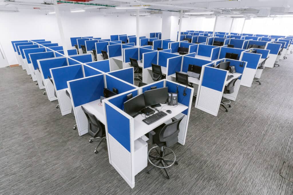 A brightly lit office space with multiple cubicles arranged in rows. Each cubicle is equipped with a computer, keyboard, phone, and headset, surrounded by blue partition walls. The area has a clean, modern look with gray carpet flooring and ergonomic chairs