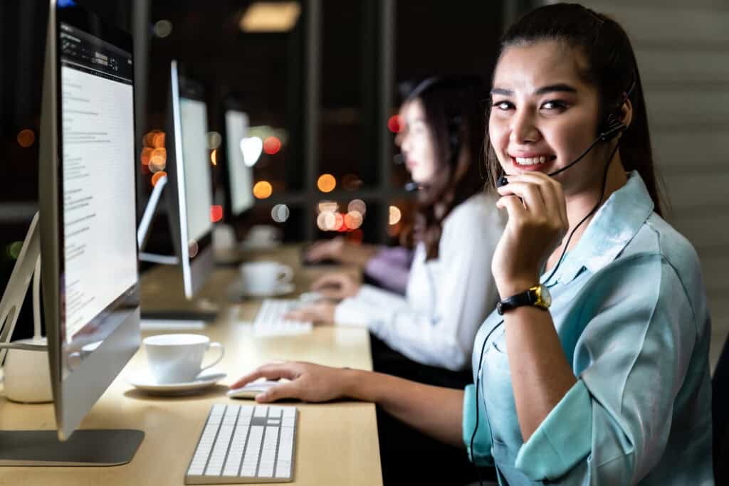 Smiling female call center agent wearing a headset while working at a computer in a modern office at night. Other agents are visible in the background, providing customer support. The image represents a professional seat leasing setup for outsourced customer service operations.