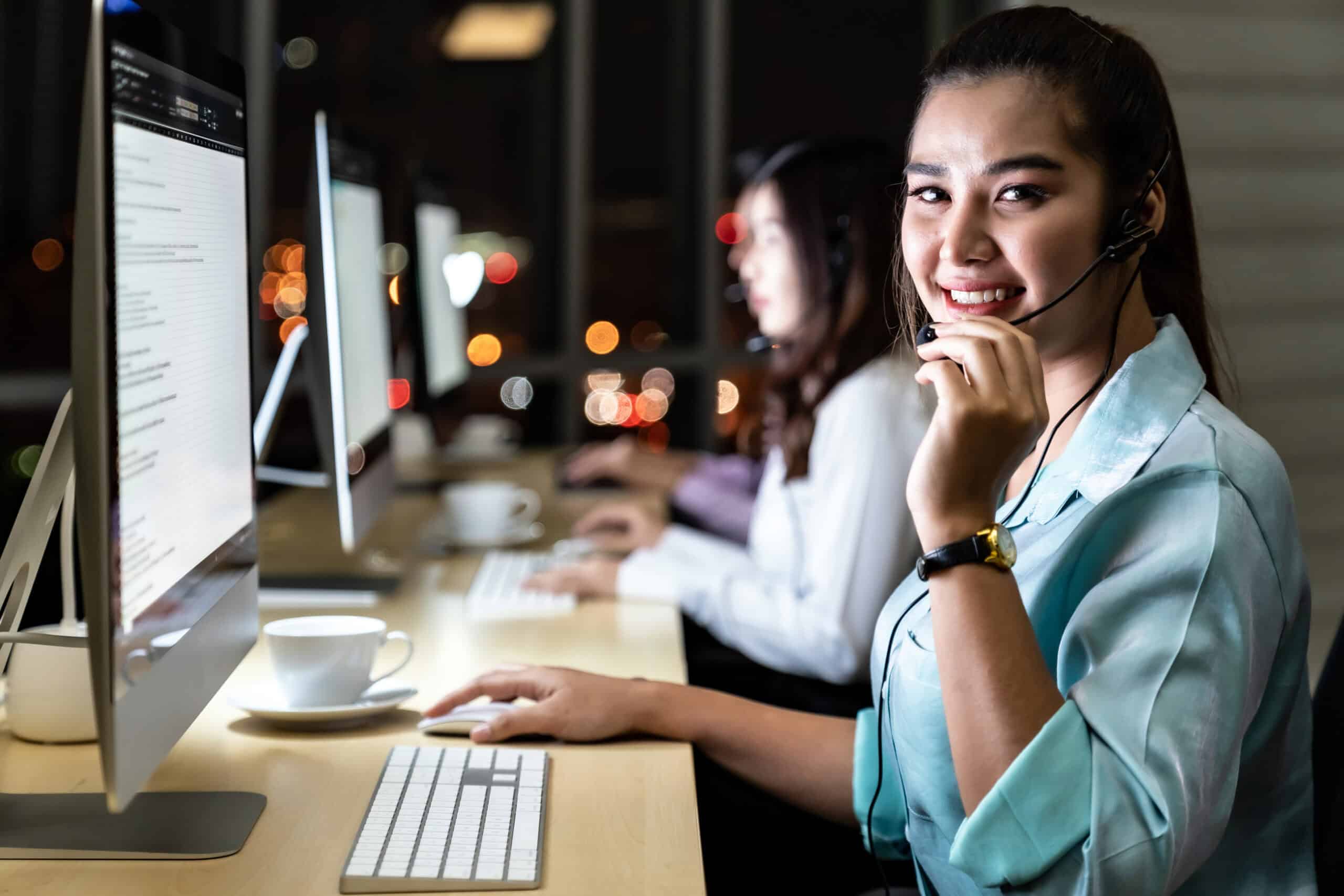 Smiling female call center agent wearing a headset while working at a computer in a modern office at night. Other agents are visible in the background, providing customer support. The image represents a professional seat leasing setup for outsourced customer service operations.