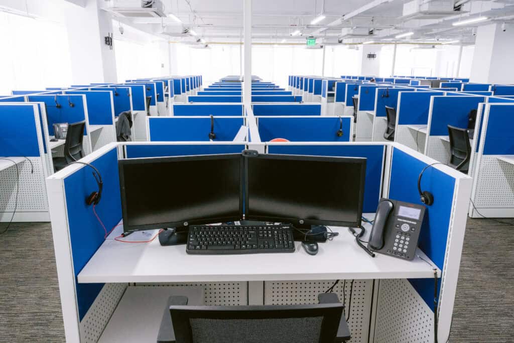 A brightly lit office space with multiple cubicles arranged in rows. Each cubicle is equipped with a computer, keyboard, phone, and headset, surrounded by blue partition walls. The area has a clean, modern look with gray carpet flooring and ergonomic chairs