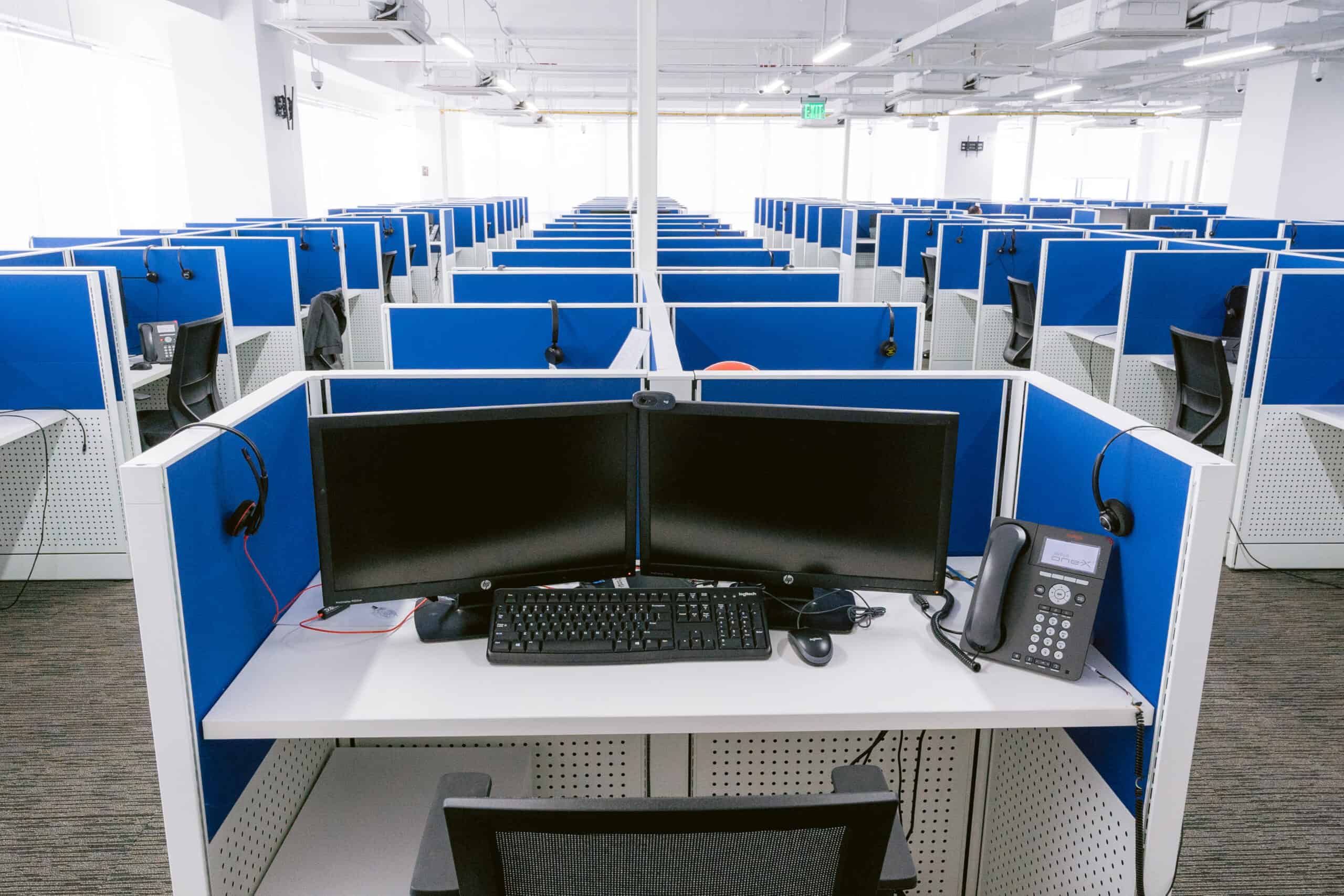 A brightly lit office space with multiple cubicles arranged in rows. Each cubicle is equipped with a computer, keyboard, phone, and headset, surrounded by blue partition walls. The area has a clean, modern look with gray carpet flooring and ergonomic chairs