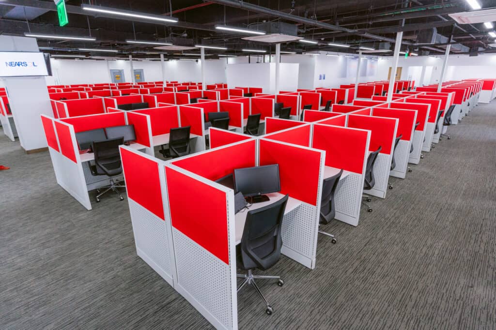 A brightly lit office space with multiple cubicles arranged in rows. Each cubicle is equipped with a computer, keyboard, phone, and headset, surrounded by red partition walls. The area has a clean, modern look with gray carpet flooring and ergonomic chairs