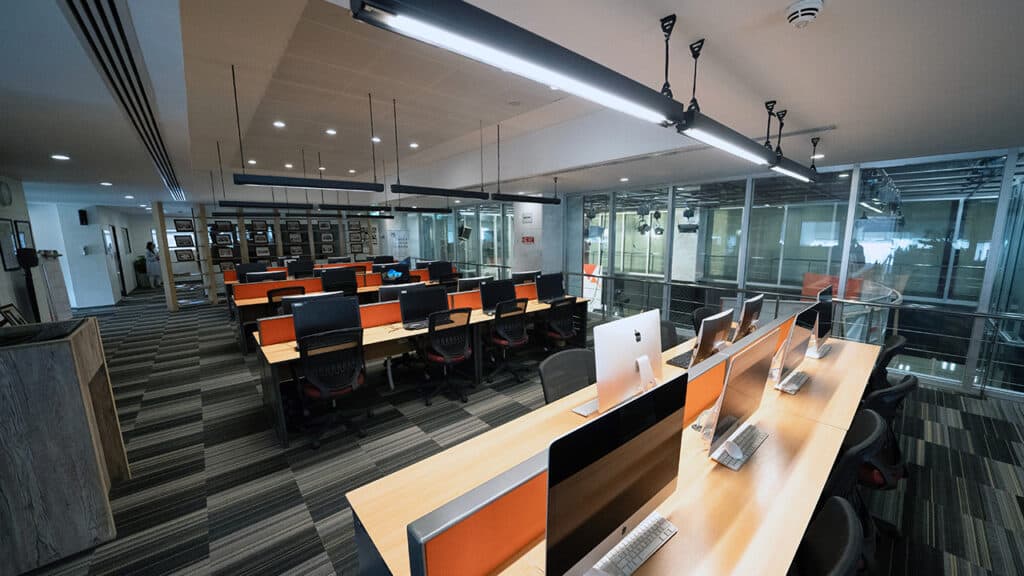 "Modern computer lab with rows of iMac desktops, ergonomic chairs, and overhead lighting in a high-tech educational or office environment, designed for digital learning or professional workspaces